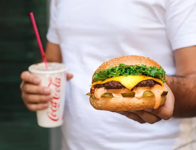 یک نفر در حال نگه داشتن یک چیزبرگر دوبل آبدار و نوشابه کوکاکولا از فست فود شیلا، آماده برای لذت بردن از یک وعده غذایی دلچسب