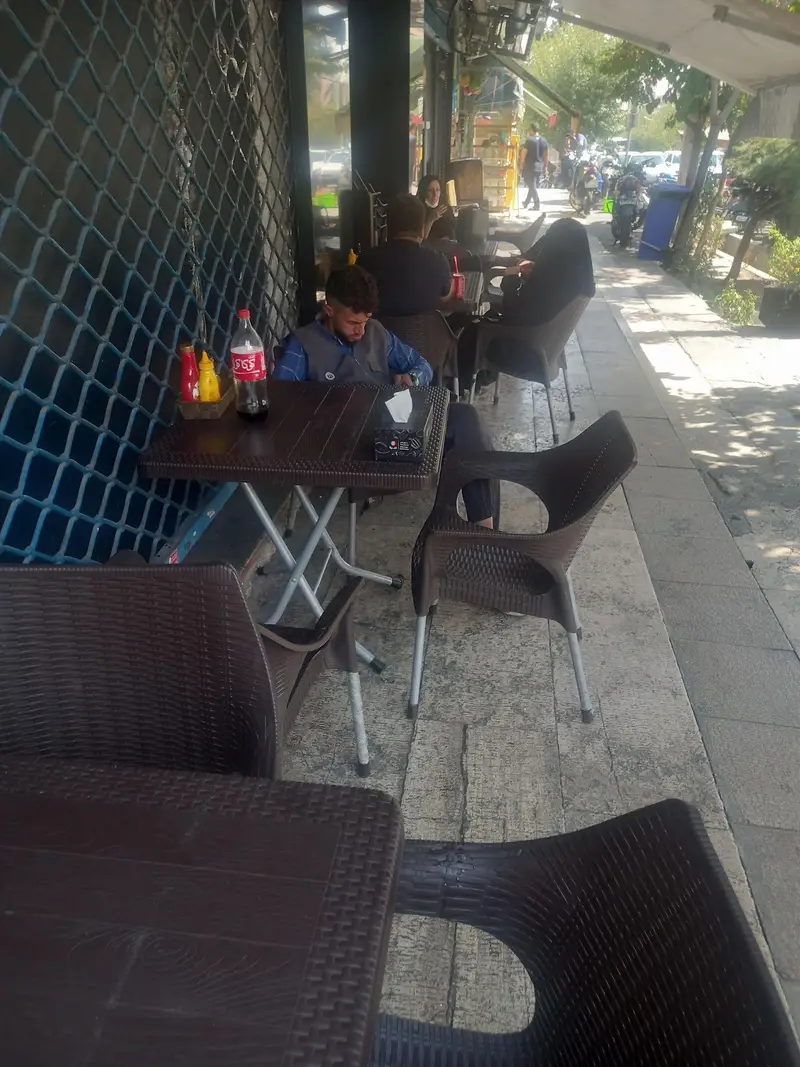 فضای باز فست فود شیلا، با میز و صندلی های حصیری قهوه ای شیک. مشتریان در حال لذت بردن از غذا و نوشیدنی با منظره خیابان.