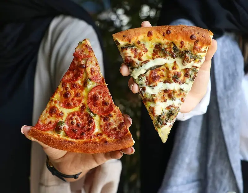 دو اسلایس پیتزای خوشمزه و تازه از فست فود شیلا، شامل پیتزا پپرونی با گوجه و فلفل و پیتزا سبزیجات با پنیر، در دست.