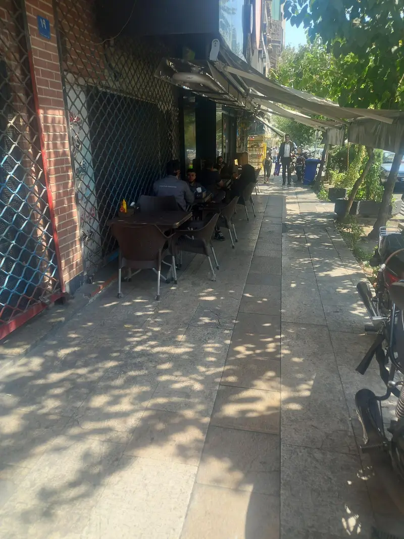 فست فود شیلا با محیط باز و دنج خود، شامل میز و صندلی برای مشتریان در حال لذت بردن از غذای خود در کنار خیابان، مناسب روزهای آفتابی.