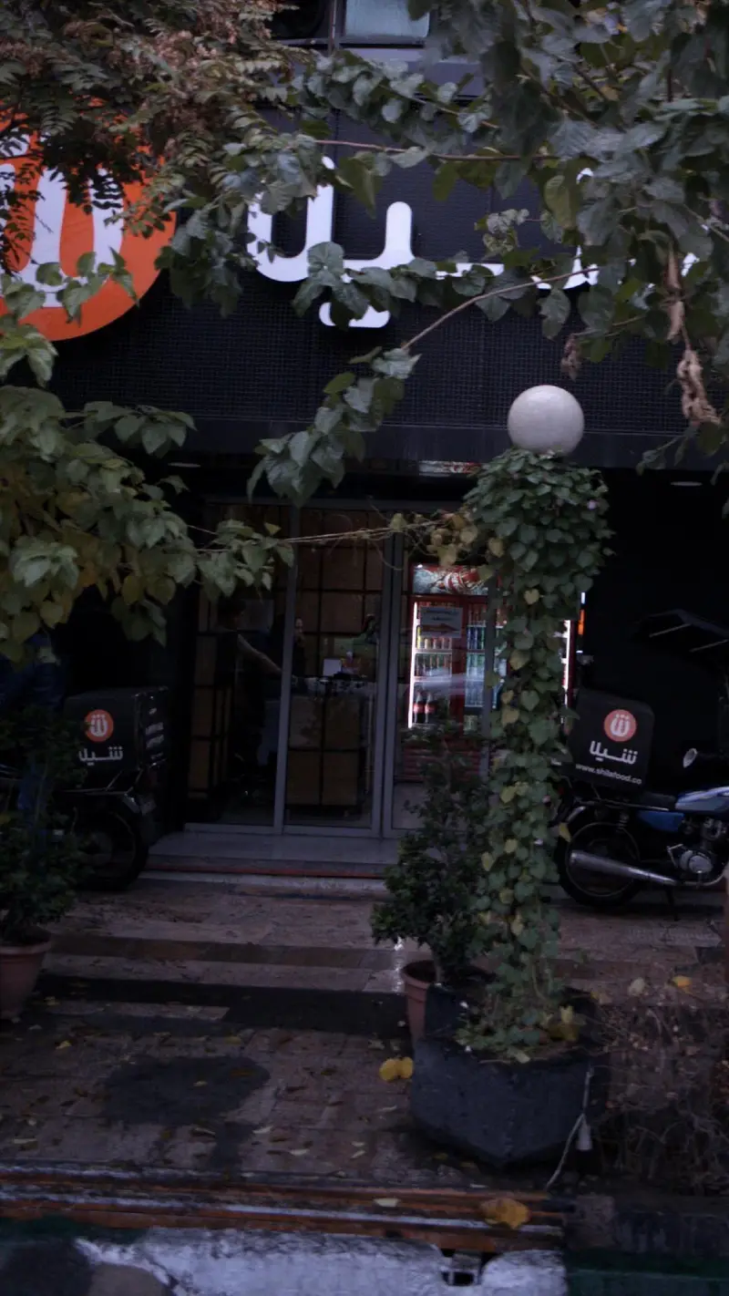 تصویر نمای بیرونی رستوران فست فود شیلا با تابلوی مشخص، ورودی و موتورسیکلت‌های دلیوری آماده ارسال سریع سفارشات آنلاین.