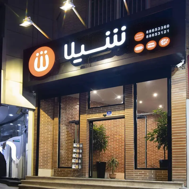 نمای بیرونی و تابلوی روشن فست فود شیلا (شعبه ونک) در شب، با شماره تلفن‌های 88653360 و 88653121 و آیکون انواع غذاها.