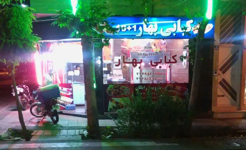 نمای بیرونی شبانه کبابی بهار با تابلوی روشن آبی و سبز، موتورسیکلت تحویل غذا در پیاده‌رو. ارائه انواع کباب و غذای بیرون‌بر.