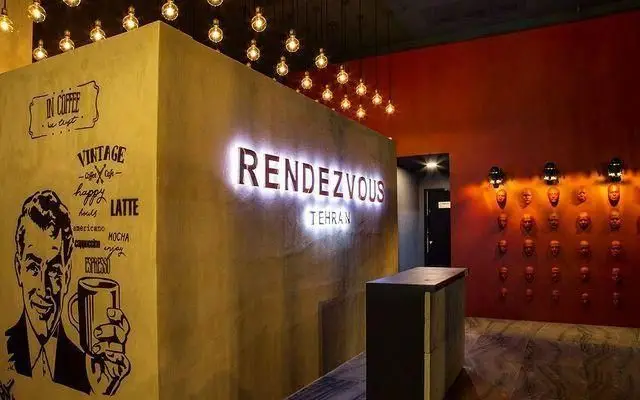 فضای داخلی کافه راندوو تهران با تابلوی روشن "RENDEZVOUS TEHRAN"، منوی نوشیدنی‌های قهوه مانند اسپرسو و لاته و طراحی هنری دیوارها.