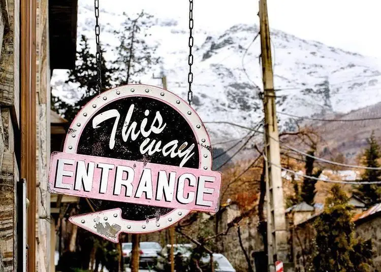 تابلو "این راه ورودی" کافه راندوو با طرح رترو صورتی و مشکی آویزان، همراه با منظره کوهستانی برفی و فضای دنج کافه.