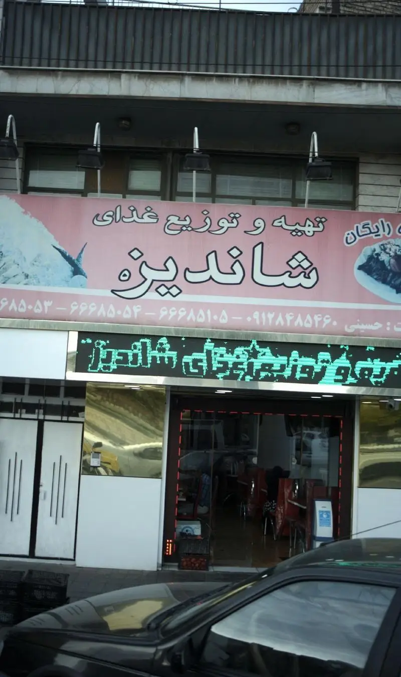 نمای بیرونی مغازه "تهیه و توزیع غذای شاندیز" با تابلوی بزرگ و مشخصات تماس برای سفارشات آنلاین و حضوری.
