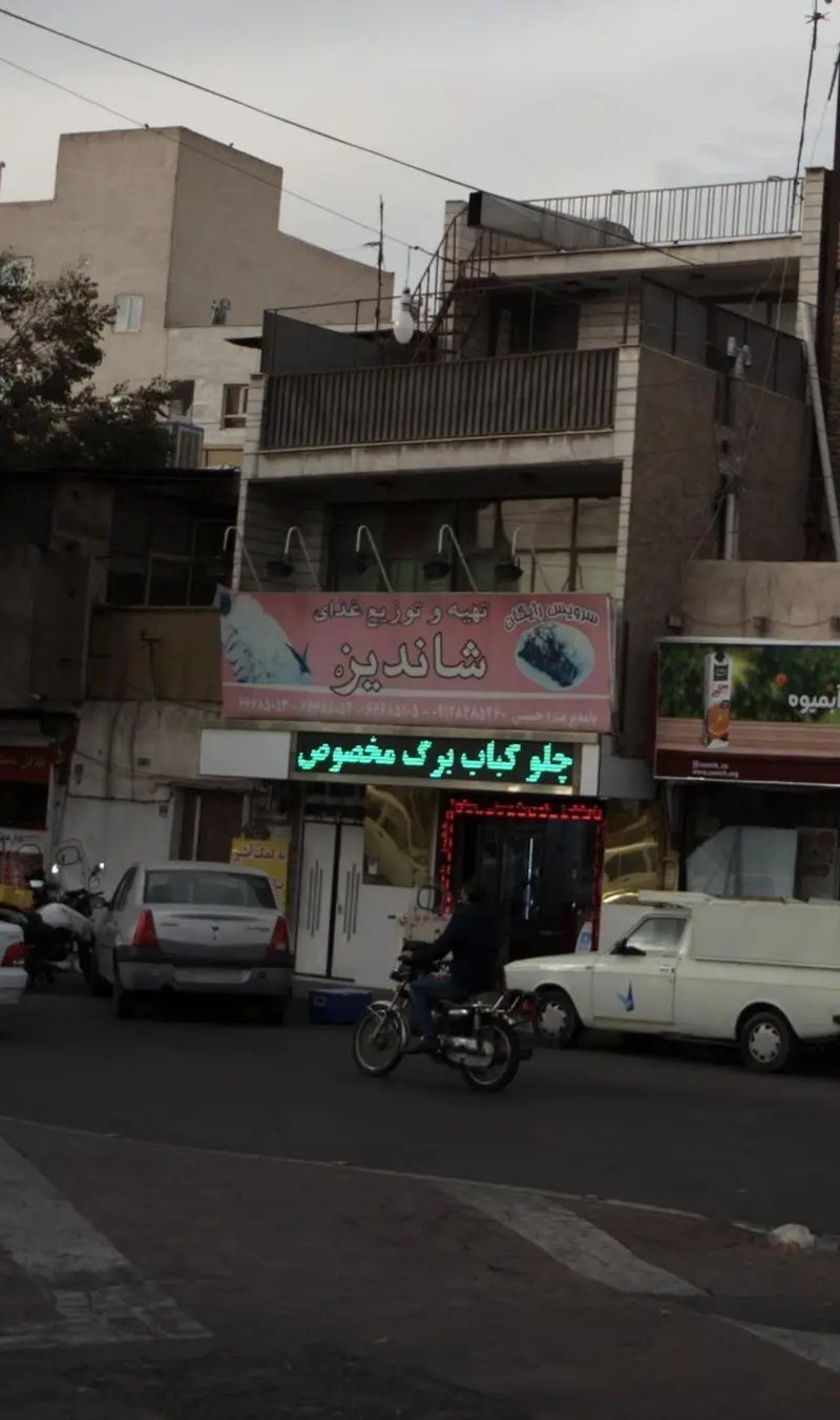 تصویری از نمای بیرونی تهیه و توزیع غذای شاندیز، همراه با تابلوی صورتی و نمایشگر 'چلو کباب برگ مخصوص' در خیابان شلوغ