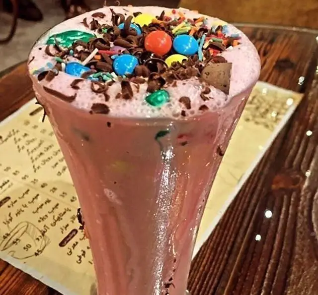 میل‌شیک صورتی خنک و جذاب با تزیین اسمارتیز رنگی، تراشه‌های شکلات و اسپرینکلز در کافه و سفره خانه تیام. دسر فانتزی.