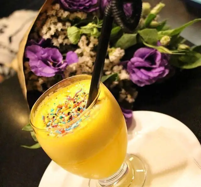 نوشیدنی خنک و خوشمزه اسموتی انبه با تزیین رنگارنگ و گل‌های بنفش در کافه تیام، مکانی دلنشین برای دورهمی شما.