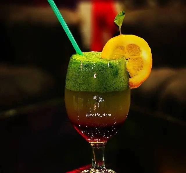 نوشیدنی خنک و گوارا با رنگ‌های سبز، کهربایی و قرمز به صورت لایه‌ای در لیوان پایه بلند، تزیین شده با لیمو و برگ نعنا، در کافه و سفره خانه تیام.