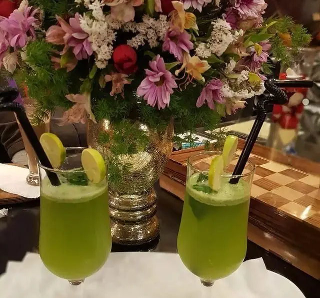 دو لیوان شربت سبز خنک با لیمو، سرو شده در کافه و سفره خانه تیام. گلدان گل زیبا و سینی چوبی شطرنجی در پس‌زمینه، محیطی آرامش‌بخش.