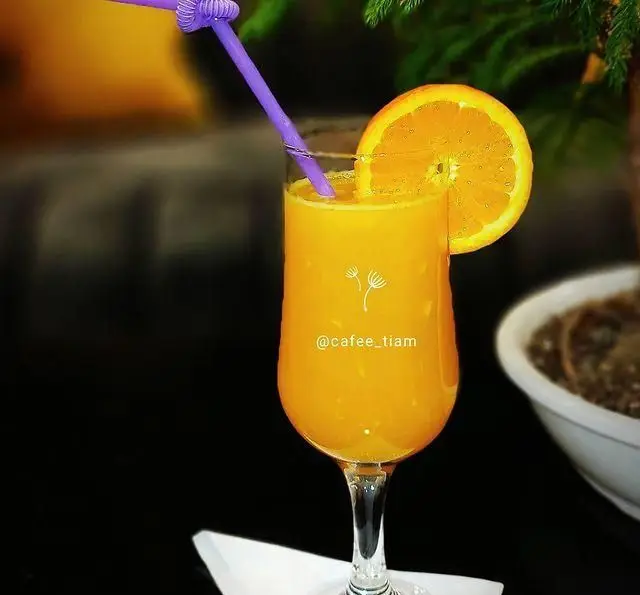 تصویر آب پرتقال تازه و طبیعی در لیوانی بلند با نی بنفش و برش پرتقال، نوشیدنی خنک و گوارا در کافه و سفره خانه تیام.