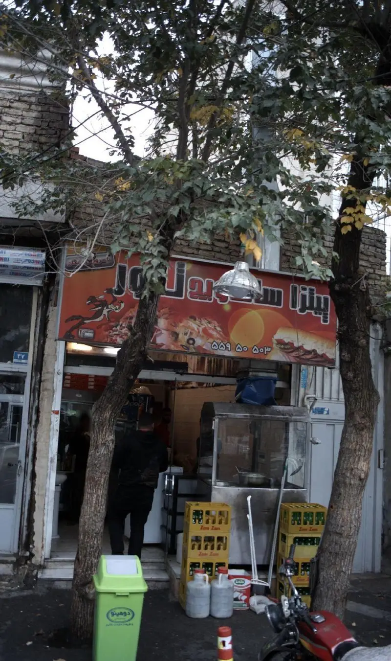 نمایی واضح از فست فود پیتزا ساندویچ تنور در خیابان، با تابلوی جذاب غذاها و مشتریان در حال ورود، همراه با درختان سبز.