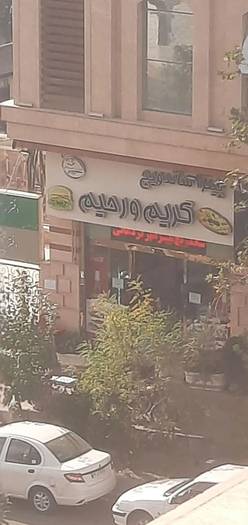 نمای بیرونی فست فود ساندویچ و پیتزا کریم و رحیم با تابلوی روشن، شامل لوگوهای همبرگر و پیتزا روی تابلو.