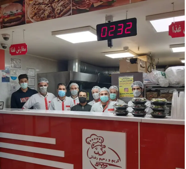 پرسنل با لبخند و ماسک در فست فود ساندویچ و پیتزا کریم و رحیم رضائی در حال آماده سازی و بسته بندی سفارشات غذا.