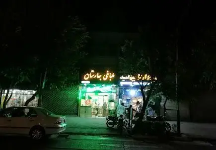 نمای بیرونی طباخی بهارستان در شب با تابلوی روشن، نورپردازی سبز داخلی و حضور مشتریان، خودرو و موتورسیکلت.