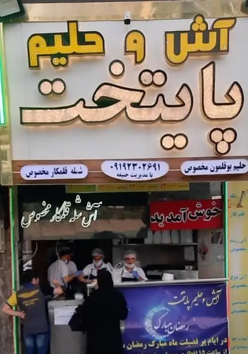 تصویری از فروشگاه آش و حلیم پایتخت با تابلوی روشن، عرضه حلیم بوقلمون و شله قلمکار مخصوص توسط کارکنان در محیطی پر جنب و جوش.