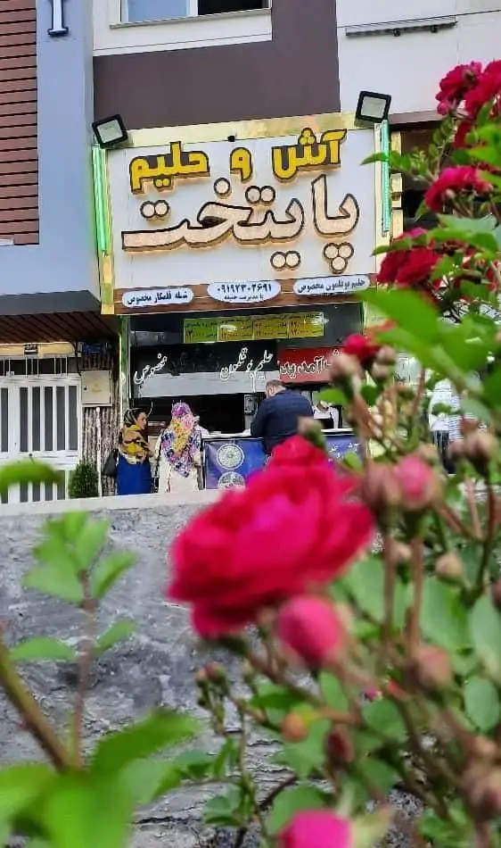 تصویری جذاب از فروشگاه آش و حلیم پایتخت، نمایش‌دهنده تابلوی روشن، مشتریان و گل‌های رز زیبا، متخصص در آش و حلیم سنتی.