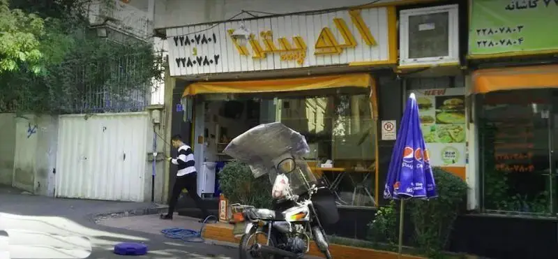 نمایی بیرونی از مغازه الو پیتزا، یک فست فود با تابلوی زرد و سفید در کنار خیابان، دارای چتر پپسی و شماره‌های تلفن روی تابلو برای تماس و سفارش.