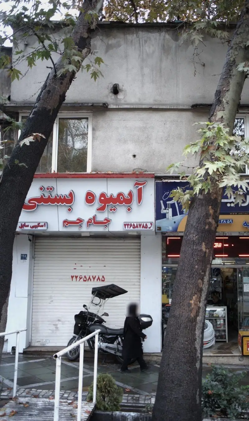 مغازه آبمیوه بستنی جام جم با تابلوی قرمز و روشن، کرکره بسته، شماره تلفن ۲۲۶۵۸۷۸۵، و درختان پاییزی در یک نمای شهری.