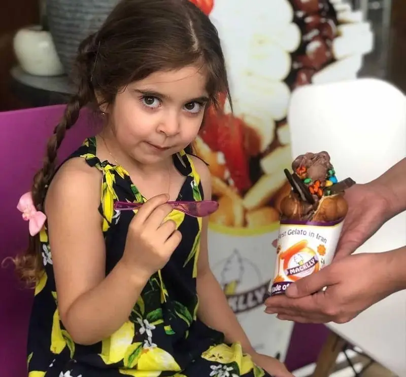 دختربچه‌ای کنجکاو با پیراهن لیمویی در بستنی مکلی نیروهوایی، ژلاتو دست‌ساز شکلاتی با تاپینگ‌های متنوع را دریافت می‌کند؛ تجربه‌ای شیرین.