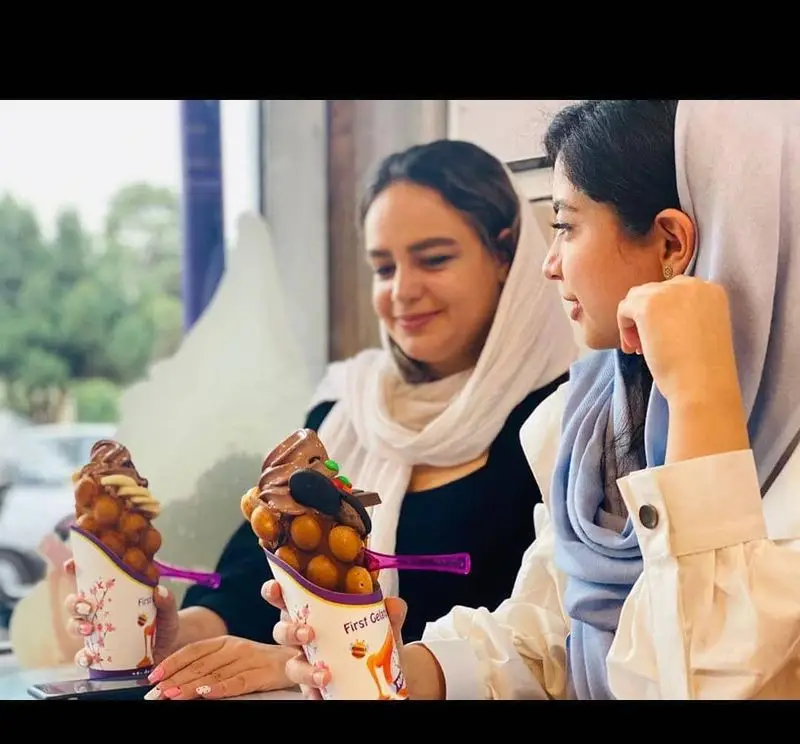 دو زن جوان در بستنی مکلی نیروهوایی با خوشحالی وافل‌های بستنی خوشمزه با تزئینات متنوع را میل می‌کنند. تجربه‌ای شیرین و دوستانه در کافه.