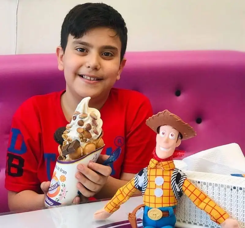 پسر بچه‌ای خندان در بستنی مکلی نیروهوایی، بستنی وافل کرم با تکه‌های شکلات، آجیل و اوریو را همراه با عروسک وودی نشان می‌دهد.