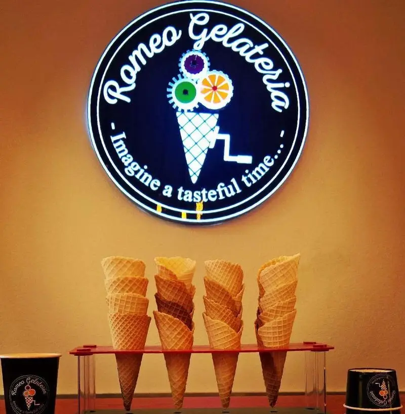 تابلوی نئونی و زیبای بستنی ایتالیایی رومئو ژلاتریا با لوگوی بستنی و چرخ‌دنده‌های رنگارنگ، در کنار مخروط‌های وافلی آماده برای سرو ژلاتوی خوشمزه.