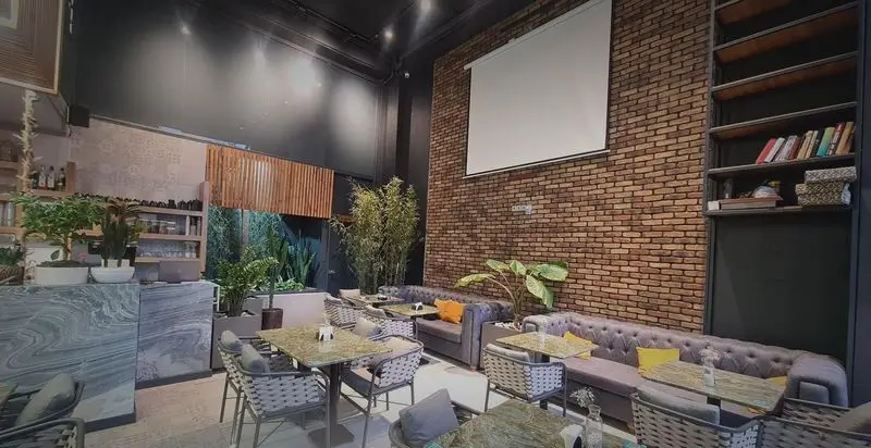 محیط دنج و مدرن کافه رستوران صدصا با دیوارهای آجری، مبلمان راحتی، گیاهان سبز، کانتر مرمری و پروژکتور برای مهمانی‌ها.