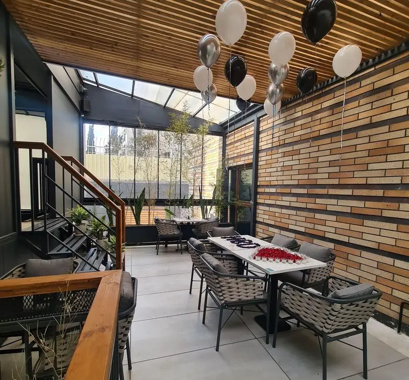 تراس دلنشین کافه رستوران صدصا با سقف چوبی و نورگیر، دیوارهای آجری، میز و صندلی‌های مدرن، بالون‌های مشکی و سفید و گیاهان بامبو