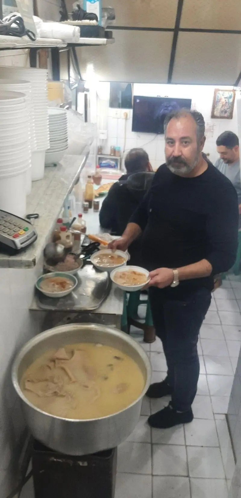 نمایی از منوچهر در طباخی پرطرفدارش، مشغول آماده‌سازی و سرو غذاهای سنتی کله پاچه در یک محیط گرم و صمیمی برای مشتریان مشتاق.