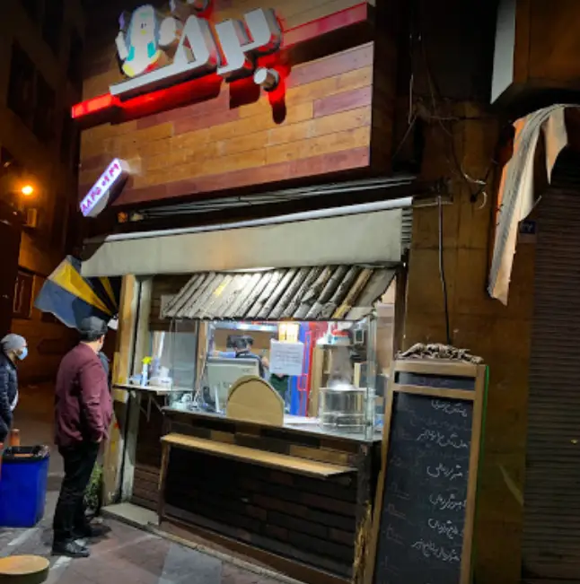 فست فود برف در شب، با تابلوی روشن، پیشخوان آماده سرویس‌دهی و مشتریانی که برای سفارش غذا منتظرند. منوی بیرون‌بر نیز دیده می‌شود.