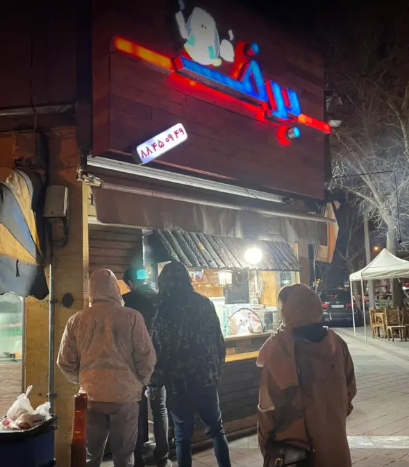 نمایی شبانه از فست فود برف با مشتریانی که در حال سفارش غذا هستند. تابلو نئون روشن و جذاب با شماره تماس 88450949.