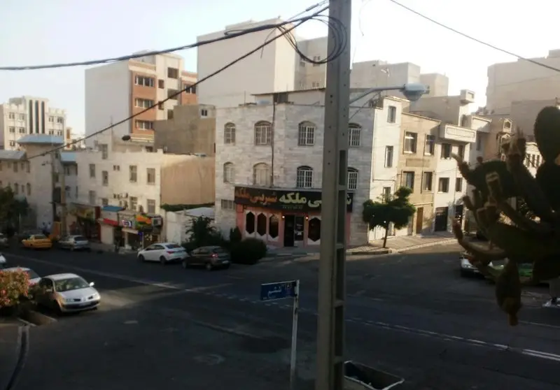 نمایی جذاب از قنادی شیرینی ملکه در محیط شهری، با ویترین زیبا، ساختمان‌های اطراف و خیابان پرتردد خودروها.