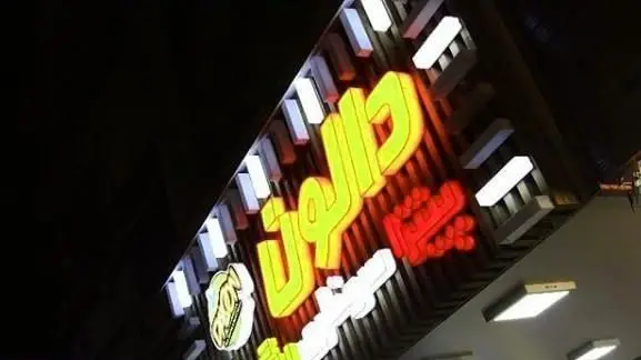 تابلوی نئونی فست فود دالون در شب، با حروف درخشان زرد و قرمز و لوگوی سفید، یک مکان پر جنب و جوش برای غذای آماده.