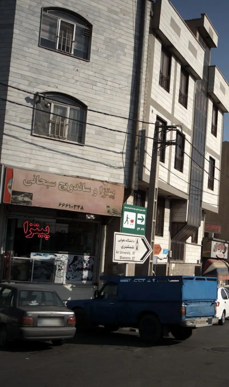 تصویری از نمای جلویی پیتزا و ساندویچ سبحانی، رستورانی محبوب در تقاطع خیابان دانشگاه هوایی و شمشیری، تهران، با تابلوی نئون پیتزا و منظره شهری.