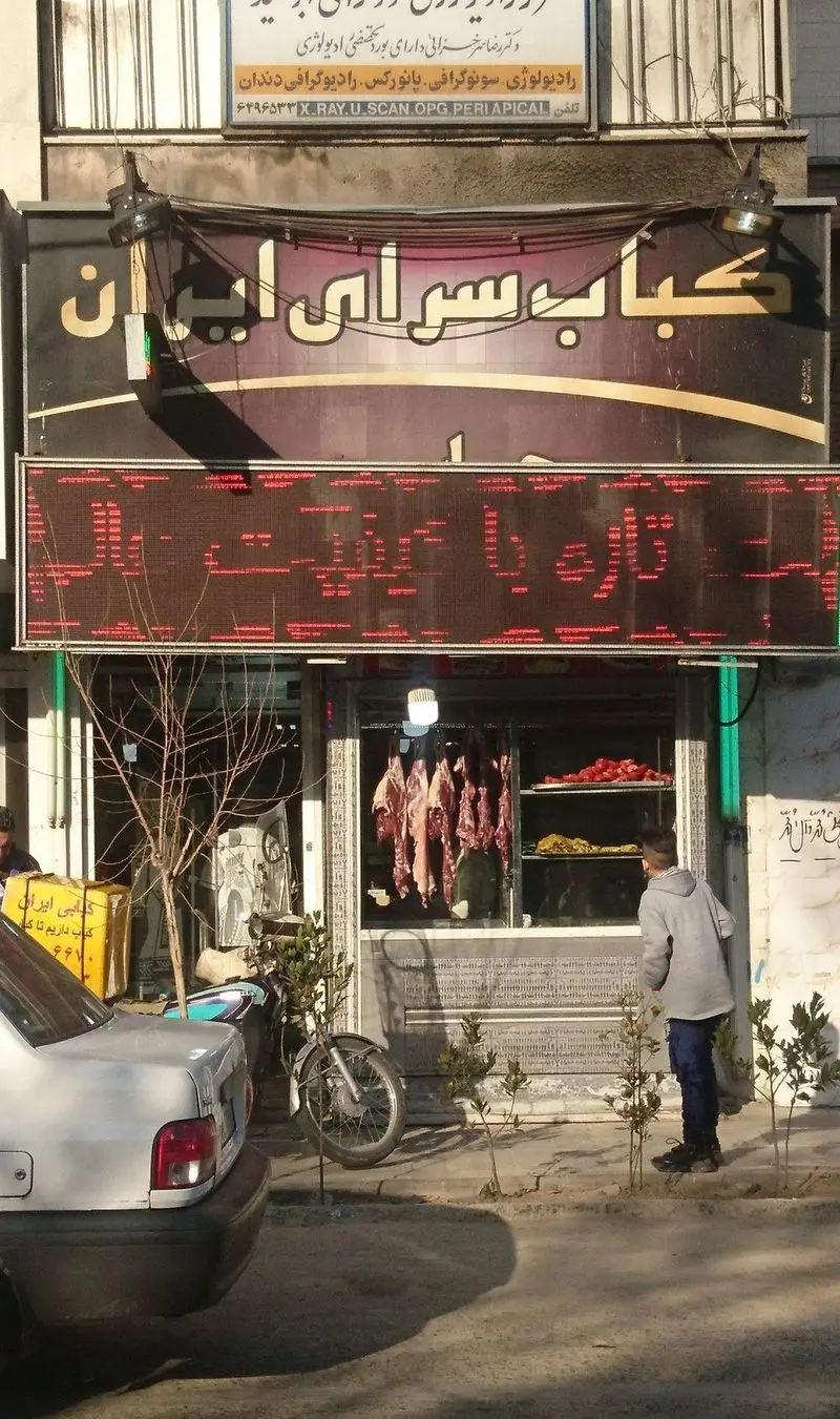 کباب سرای ایران: نمای بیرونی مغازه کباب فروشی با گوشت تازه آویزان، تابلوی اصلی کباب سرای ایران و تابلوی LED "تازه با کیفیت" در شهر.