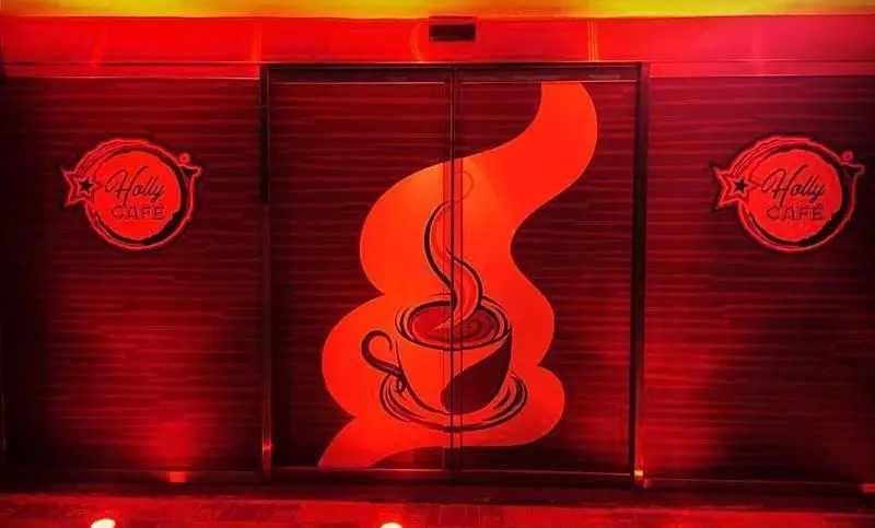 تصویر ورودی شیک و مدرن هالی کافه با نورپردازی گرم و قرمز، لوگوهای برجسته Holly Cafe و طرح بزرگ و هنری فنجان قهوه داغ روی درب‌ها.
