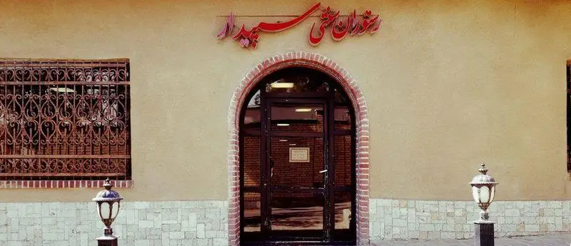 ورودی دنج و سنتی رستوران سپیدار، نمایی از معماری ایرانی با خطاطی زیبا و چراغ‌های قدیمی، مکانی دلنشین برای صرف غذا.