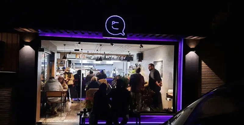 نمای خارجی شبانه رستوران کوچینا پیتزا با لوگوی نئون بنفش و مشتریان داخل، نورپردازی جذاب و فضای گرم و دلنشین برای صرف پیتزا.