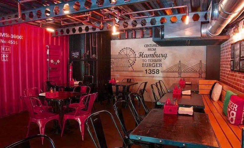 دکوراسیون داخلی منحصر به فرد رستوران برگر ۱۳۵۸ در تهران با تم صنعتی، دیوارهای کانتینری قرمز، میز و صندلی مدرن و نورپردازی گرم.
