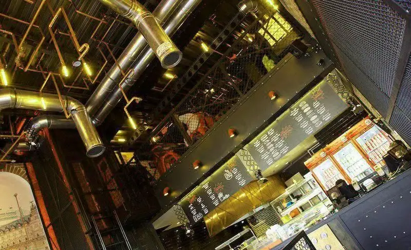 نمای داخلی فست فود برگر ۱۳۵۸ با دکوراسیون خاص صنعتی، لوله‌های فلزی روی سقف، طبقه دوم مشبک و پیشخوان سفارش.