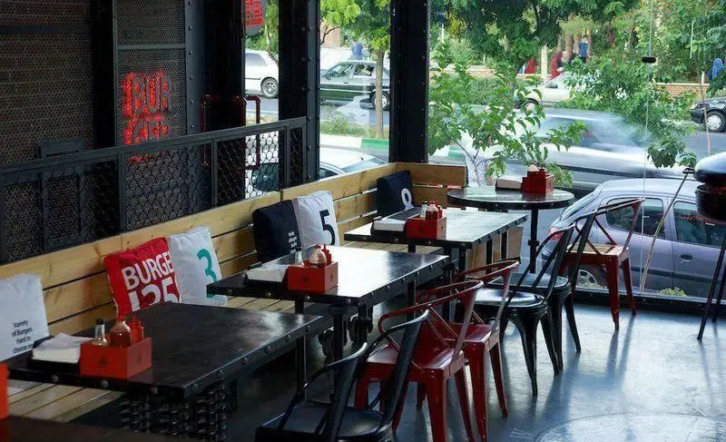 فضای داخلی دنج و مدرن رستوران برگر ۱۳۵۸ با میزهای تیره، صندلی‌های فلزی رنگی، کوسن‌های برند و منظره خیابان از پنجره بزرگ