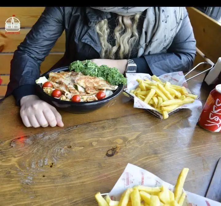 نمایی از مشتری در فست فود برگر ۱۳۵۸ در حال لذت بردن از سالاد مرغ گریل شده، سیب زمینی سرخ کرده و نوشابه روی میز چوبی.