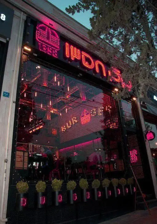 نمای بیرونی جذاب رستوران برگر ۱۳۵۸ با نورپردازی نئون صورتی و لوگوی تاج، انعکاس محیط داخلی مدرن و شیک برای فست فود.