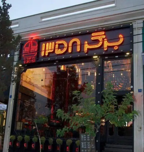 تصویر نمای خارجی کسب و کار برگر ۱۳۵۸، یک رستوران فست فود با تابلوی نئونی نارنجی رنگ و لوگوی برگر روشن. ویترین شیشه ای بزرگ آن بازتاب نور محیط را نشان می دهد.