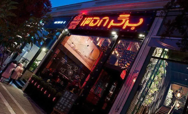 نمای بیرونی جذاب رستوران فست فود برگر ۱۳۵۸ در شب، با تابلوی نئون روشن و طراحی مدرن شیشه‌ای، جلب توجه رهگذران.