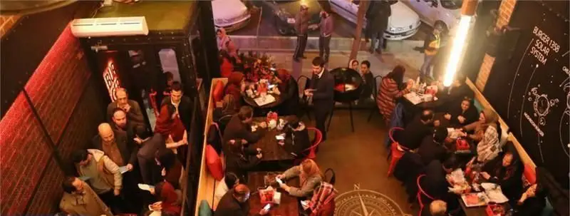 نمایی از فضای داخلی رستوران شلوغ برگر ۱۳۵۸ با مشتریان در حال صرف غذا و دکوراسیون مدرن شامل دیوار طرح منظومه شمسی و نورپردازی جذاب.