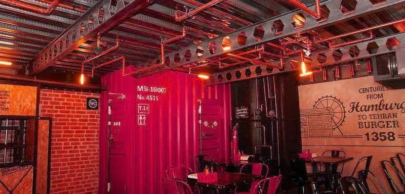 فضای داخلی خاص و صنعتی رستوران برگر ۱۳۵۸ با دیوارهای کانتینری قرمز، سقف فلزی اکسپوز، لوله‌های مسی و نورپردازی جذاب.