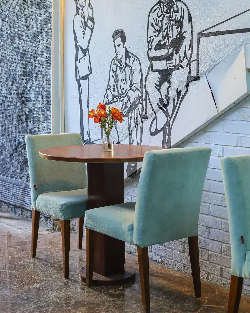 دکوراسیون داخلی کافه بینج: میز گرد چوبی و صندلی‌های راحت سبز روشن، دیوارنگاره هنری چشم‌نواز و گلدان گل‌های نارنجی روی میز.